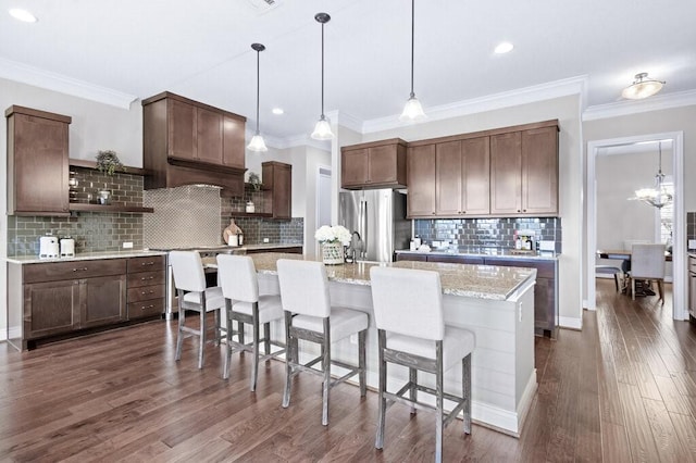 kitchen featuring pendant lighting, stainless steel fridge, dark brown cabinets, light stone countertops, and an island with sink