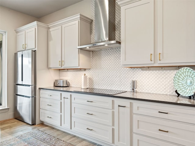 kitchen with backsplash, black electric stovetop, white cabinets, wall chimney range hood, and stainless steel refrigerator