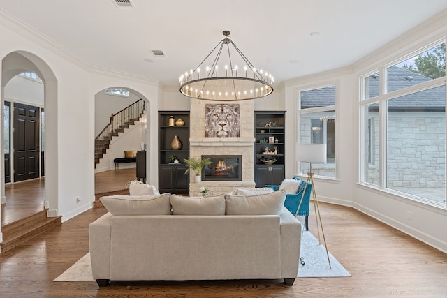 living room with built in shelves, a stone fireplace, hardwood / wood-style floors, a chandelier, and ornamental molding