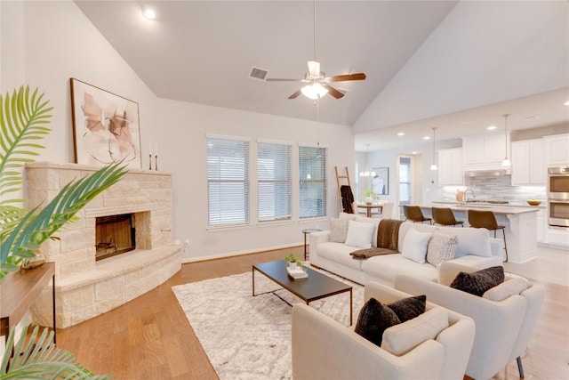 living room featuring ceiling fan, a stone fireplace, high vaulted ceiling, and light hardwood / wood-style flooring