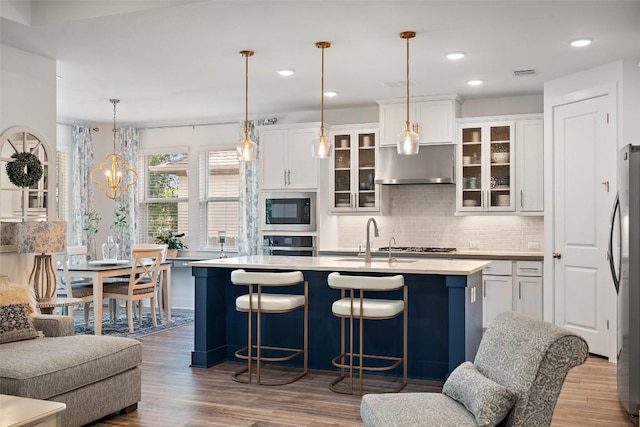 kitchen featuring built in microwave, hanging light fixtures, ventilation hood, oven, and a kitchen island with sink