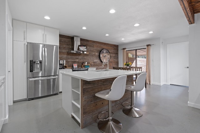 kitchen featuring wall chimney exhaust hood, wooden walls, a center island with sink, stainless steel fridge with ice dispenser, and white cabinetry