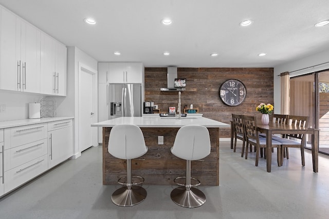 kitchen featuring a kitchen breakfast bar, stainless steel fridge with ice dispenser, wood walls, a kitchen island with sink, and white cabinets