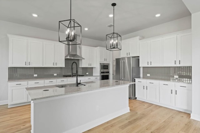 kitchen featuring a center island with sink, appliances with stainless steel finishes, wall chimney range hood, and white cabinets