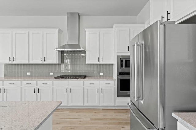 kitchen featuring wall chimney range hood, appliances with stainless steel finishes, white cabinetry, backsplash, and light stone countertops