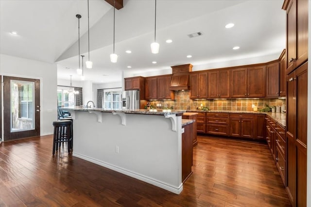 kitchen featuring a breakfast bar, hanging light fixtures, stainless steel refrigerator with ice dispenser, an island with sink, and light stone counters