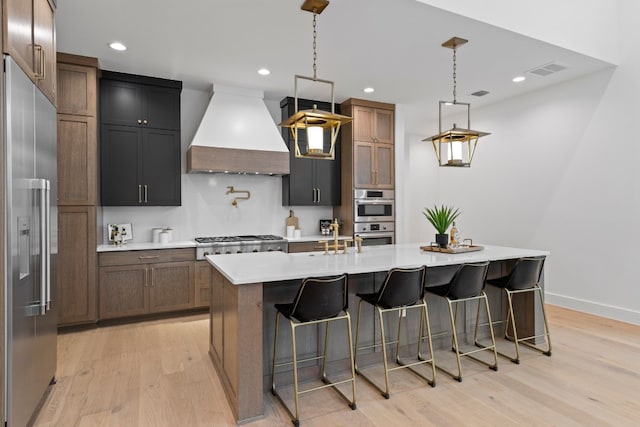 kitchen featuring light wood-type flooring, custom range hood, an island with sink, and appliances with stainless steel finishes