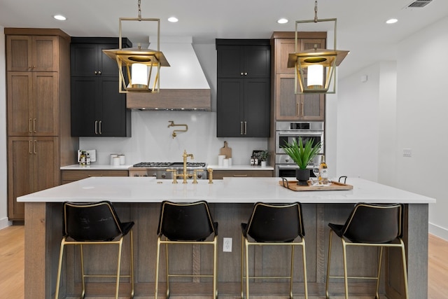 kitchen featuring double oven, light hardwood / wood-style floors, premium range hood, and a large island with sink