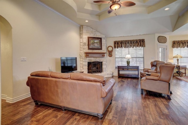 living room featuring ornamental molding, a raised ceiling, ceiling fan, dark wood-type flooring, and a stone fireplace