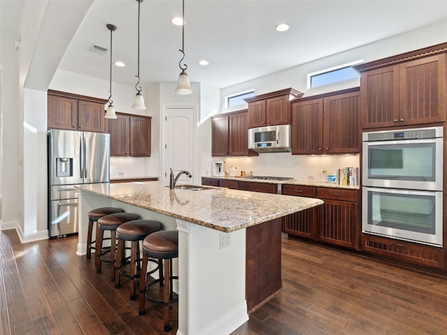 kitchen featuring a center island with sink, sink, hanging light fixtures, light stone countertops, and appliances with stainless steel finishes