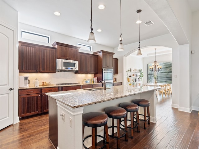 kitchen with light stone countertops, stainless steel appliances, sink, pendant lighting, and an island with sink