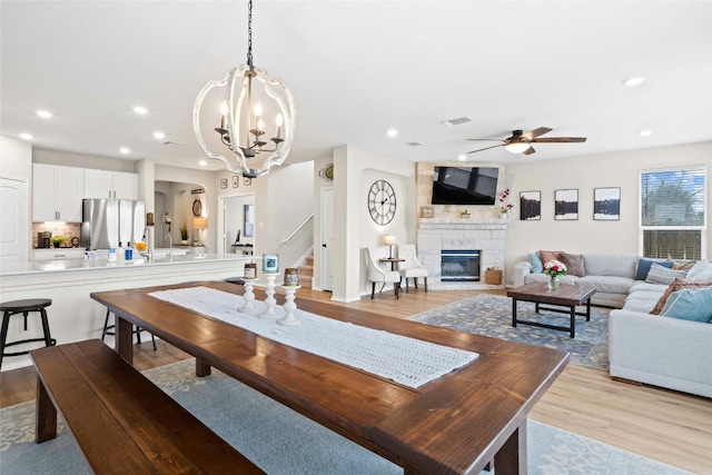 dining space featuring a stone fireplace, light hardwood / wood-style floors, and ceiling fan with notable chandelier