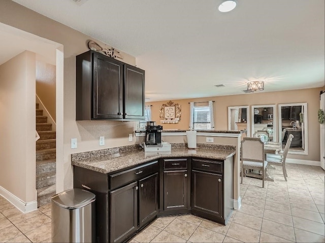 kitchen with kitchen peninsula, light tile patterned floors, dark brown cabinetry, and dark stone counters