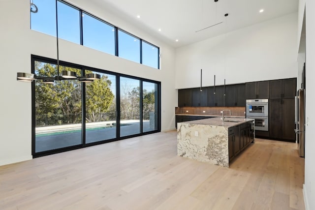 kitchen featuring light stone counters, stainless steel double oven, a high ceiling, light hardwood / wood-style floors, and an island with sink