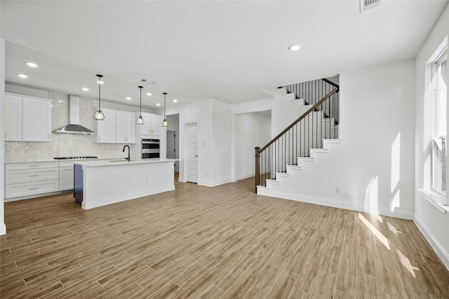 kitchen with white cabinets, wall chimney range hood, sink, an island with sink, and decorative light fixtures