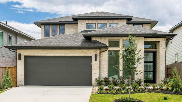 prairie-style home with fence, driveway, a shingled roof, an attached garage, and brick siding