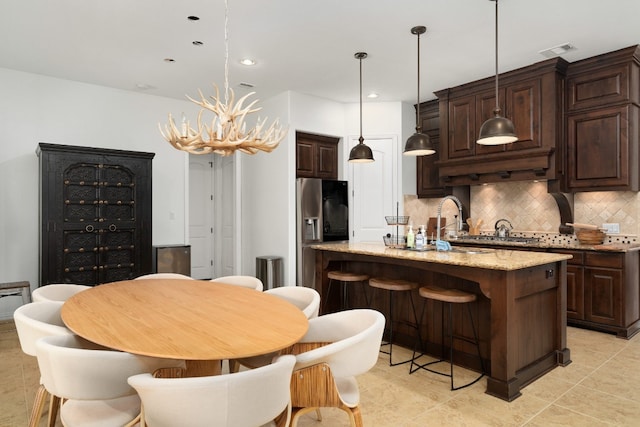 kitchen featuring stainless steel fridge, an island with sink, pendant lighting, light stone countertops, and backsplash