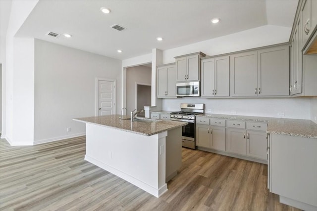 kitchen with sink, light stone counters, gray cabinets, a center island with sink, and appliances with stainless steel finishes