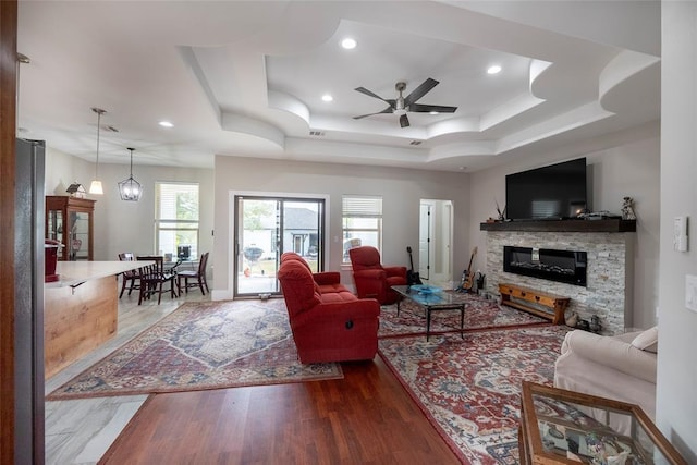 living room featuring hardwood / wood-style flooring, ceiling fan with notable chandelier, a stone fireplace, and a raised ceiling