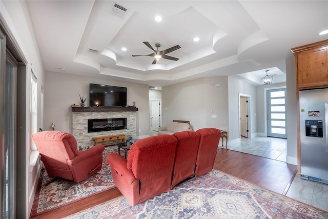 living room with ceiling fan with notable chandelier, light hardwood / wood-style floors, a raised ceiling, and a stone fireplace