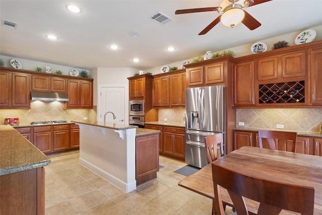 kitchen featuring appliances with stainless steel finishes, tasteful backsplash, light stone counters, a kitchen island with sink, and ceiling fan