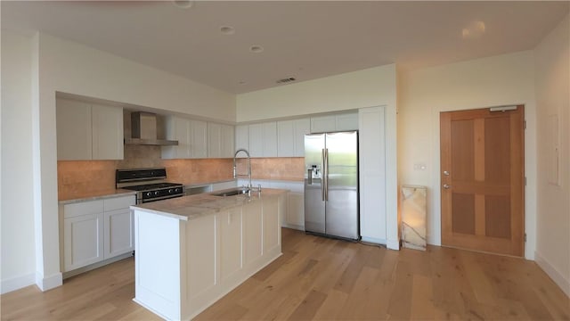 kitchen with stove, a kitchen island with sink, wall chimney range hood, sink, and stainless steel refrigerator with ice dispenser