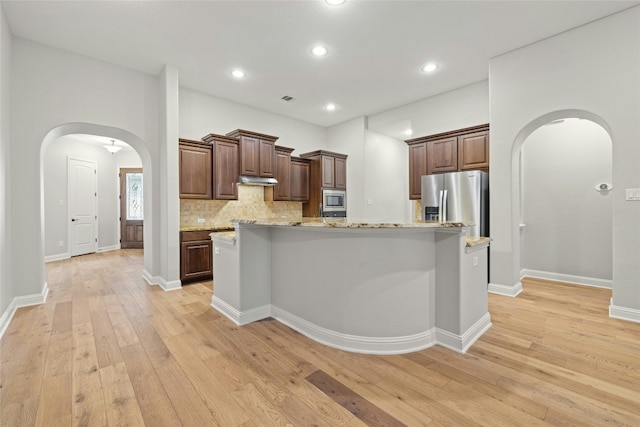 kitchen featuring appliances with stainless steel finishes, an island with sink, decorative backsplash, light stone countertops, and light wood-type flooring