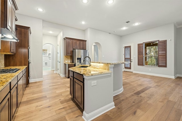 kitchen featuring light stone counters, stainless steel fridge with ice dispenser, a center island with sink, light wood-type flooring, and backsplash