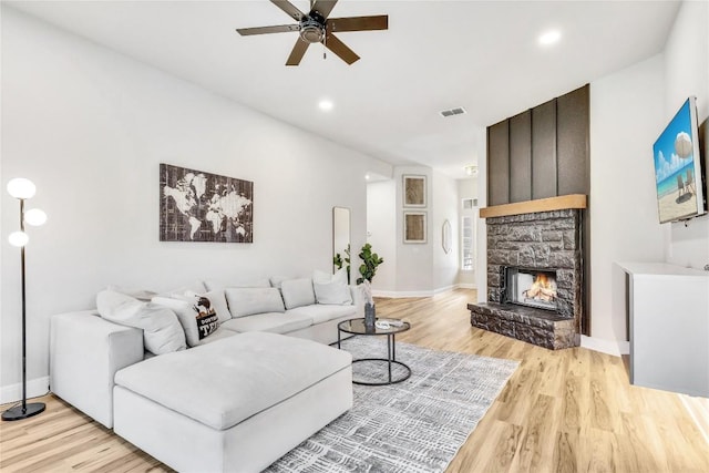 living area with visible vents, ceiling fan, a stone fireplace, light wood-type flooring, and baseboards