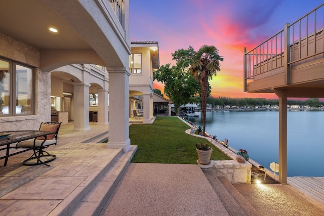 patio terrace at dusk with a lawn, a water view, a balcony, and a dock