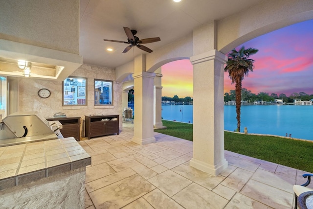 patio terrace at dusk featuring an outdoor kitchen, a water view, ceiling fan, and a lawn