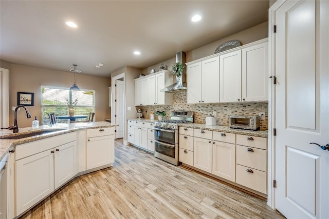 kitchen with range with two ovens, wall chimney range hood, sink, hanging light fixtures, and white cabinetry