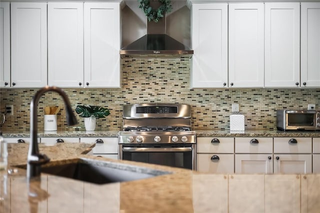 kitchen with white cabinets, wall chimney exhaust hood, stainless steel gas range oven, and backsplash