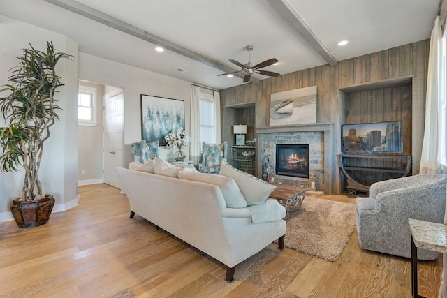 living room with beam ceiling, a stone fireplace, ceiling fan, and light hardwood / wood-style floors