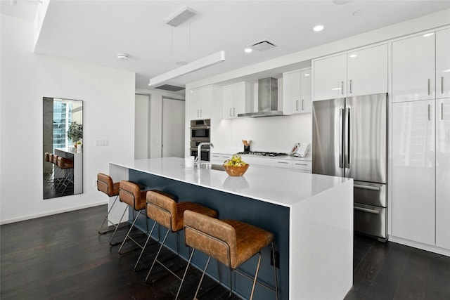 kitchen featuring a kitchen breakfast bar, wall chimney exhaust hood, stainless steel appliances, a center island with sink, and white cabinetry