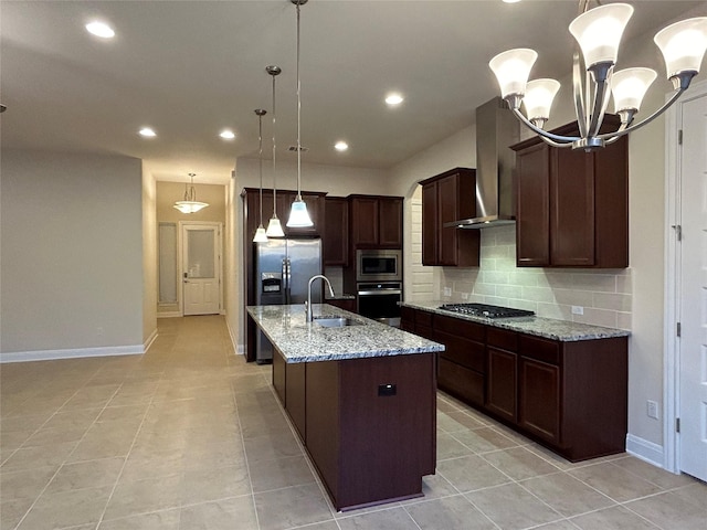 kitchen featuring wall chimney exhaust hood, light stone counters, pendant lighting, a kitchen island with sink, and appliances with stainless steel finishes