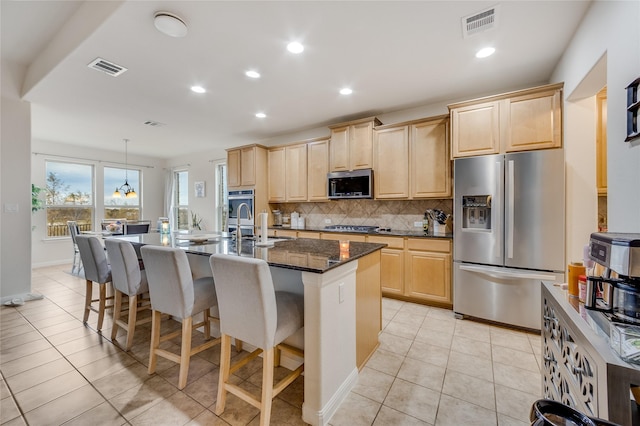 kitchen with light brown cabinets, stainless steel appliances, tasteful backsplash, dark stone countertops, and a center island with sink