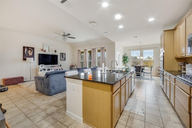 kitchen featuring a center island with sink, ceiling fan, dark stone countertops, light brown cabinetry, and sink