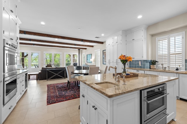 kitchen featuring sink, stainless steel double oven, beamed ceiling, an island with sink, and white cabinets