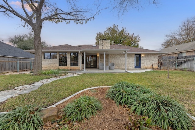 rear view of house featuring a patio, stone siding, a chimney, fence, and a yard