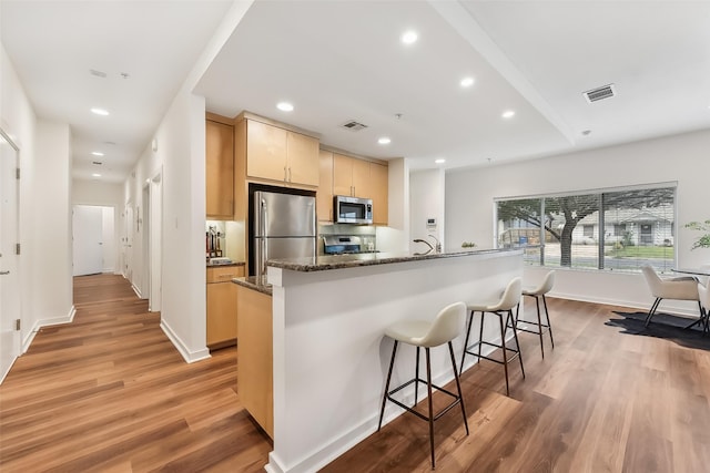 kitchen featuring hardwood / wood-style floors, dark stone counters, light brown cabinetry, appliances with stainless steel finishes, and a breakfast bar area