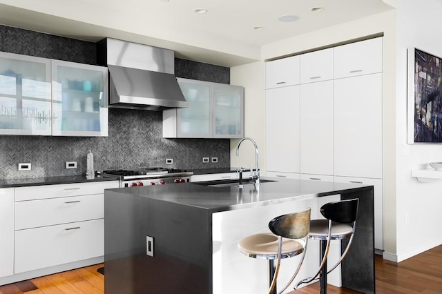 kitchen featuring white cabinetry, sink, wall chimney exhaust hood, a kitchen island with sink, and range