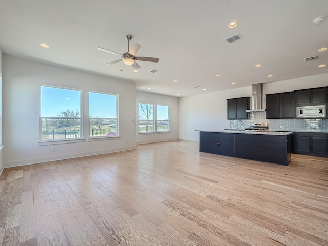 kitchen featuring a kitchen island with sink, light hardwood / wood-style floors, stainless steel appliances, light stone counters, and wall chimney range hood