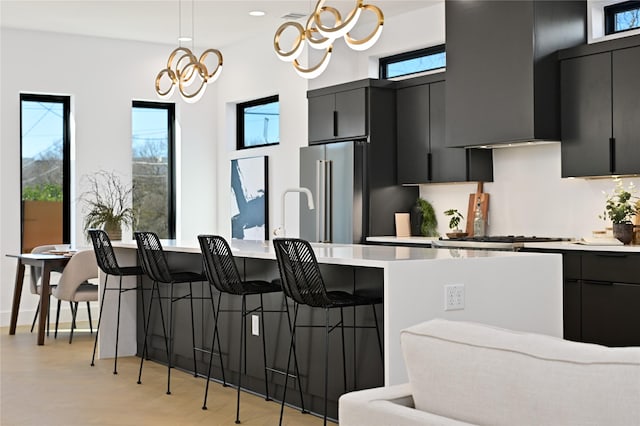 kitchen featuring high end refrigerator, an inviting chandelier, wall chimney exhaust hood, decorative light fixtures, and a kitchen bar