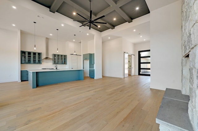 kitchen with a towering ceiling, wall chimney exhaust hood, ceiling fan, a large island with sink, and decorative light fixtures