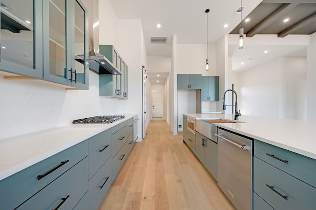 kitchen featuring light stone countertops, wall chimney exhaust hood, hanging light fixtures, stainless steel appliances, and light wood-type flooring