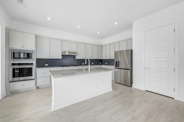 kitchen featuring sink, a kitchen island with sink, stainless steel appliances, light stone counters, and decorative backsplash