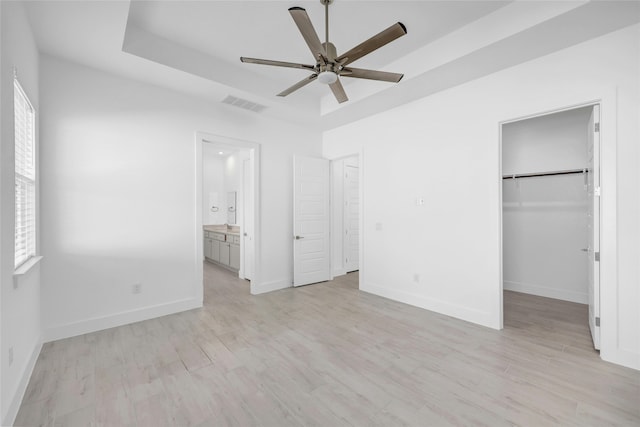 unfurnished bedroom featuring ceiling fan, light wood-type flooring, a spacious closet, connected bathroom, and a tray ceiling