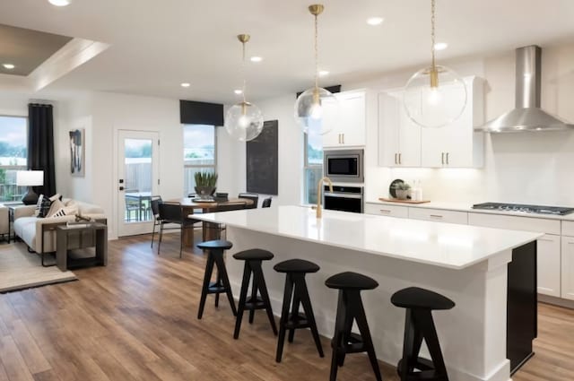 kitchen with appliances with stainless steel finishes, hanging light fixtures, an island with sink, white cabinets, and wall chimney range hood