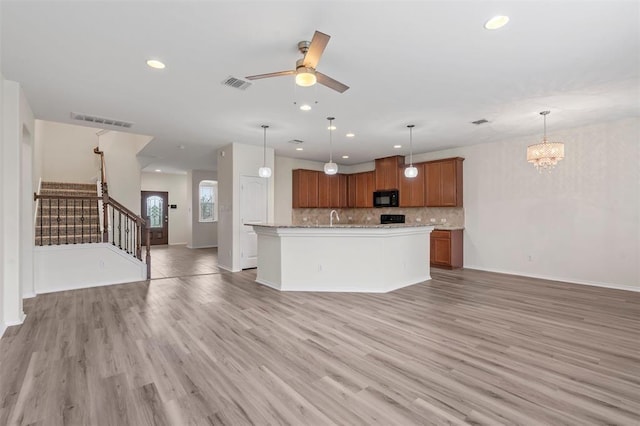 kitchen with pendant lighting, ceiling fan with notable chandelier, light hardwood / wood-style flooring, decorative backsplash, and an island with sink